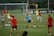 A group of female soccer players engaged in a practice session on a grass field. They wear red and blue uniforms, with some in training bibs. A visible goalpost, cones, and soccer balls are scattered around. In the background, a few spectators watch from sidelines near a banner with logos.