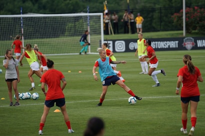 A group of female soccer players engaged in a practice session on a grass field. They wear red and blue uniforms, with some in training bibs. A visible goalpost, cones, and soccer balls are scattered around. In the background, a few spectators watch from sidelines near a banner with logos.