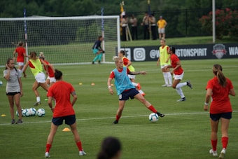A group of female soccer players engaged in a practice session on a grass field. They wear red and blue uniforms, with some in training bibs. A visible goalpost, cones, and soccer balls are scattered around. In the background, a few spectators watch from sidelines near a banner with logos.