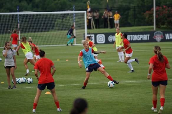 A group of female soccer players engaged in a practice session on a grass field. They wear red and blue uniforms, with some in training bibs. A visible goalpost, cones, and soccer balls are scattered around. In the background, a few spectators watch from sidelines near a banner with logos.