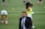 A man in a suit and tie stands on a grassy sports field. In the background, several athletes are warming up, wearing athletic gear and running. The scene suggests a training or pre-game session.