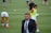 A man in a suit and tie stands on a grassy sports field. In the background, several athletes are warming up, wearing athletic gear and running. The scene suggests a training or pre-game session.