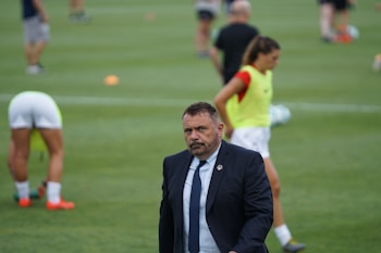 A man in a suit and tie stands on a grassy sports field. In the background, several athletes are warming up, wearing athletic gear and running. The scene suggests a training or pre-game session.