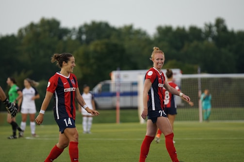 Two soccer players wearing red and blue uniforms are on a field, smiling and walking. There are other players and a goalpost in the background, with a lush, green backdrop and overcast sky.