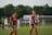 Two soccer players wearing red and blue uniforms are on a field, smiling and walking. There are other players and a goalpost in the background, with a lush, green backdrop and overcast sky.