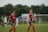 Two soccer players wearing red and blue uniforms are on a field, smiling and walking. There are other players and a goalpost in the background, with a lush, green backdrop and overcast sky.