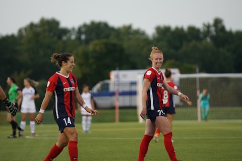 Two soccer players wearing red and blue uniforms are on a field, smiling and walking. There are other players and a goalpost in the background, with a lush, green backdrop and overcast sky.