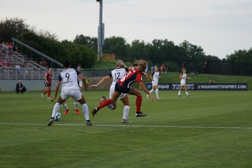 A competitive women's soccer match taking place on a grassy field. Players in red and black are attempting to gain possession of the ball, while players in white are defending. The scene conveys action and movement, with players focused on the game. Spectators are seated in the background, and there are advertisements along the sidelines.