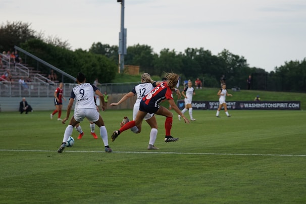 A competitive women's soccer match taking place on a grassy field. Players in red and black are attempting to gain possession of the ball, while players in white are defending. The scene conveys action and movement, with players focused on the game. Spectators are seated in the background, and there are advertisements along the sidelines.