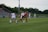 A competitive women's soccer match taking place on a grassy field. Players in red and black are attempting to gain possession of the ball, while players in white are defending. The scene conveys action and movement, with players focused on the game. Spectators are seated in the background, and there are advertisements along the sidelines.