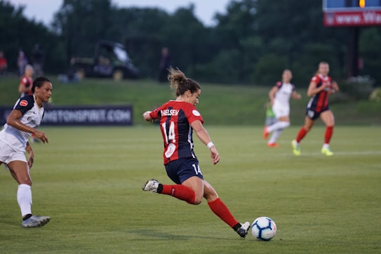 A soccer match in action with players in motion on a grass field. One player is preparing to kick the ball, while others anticipate movement. The scene includes a background of green grass and trees.