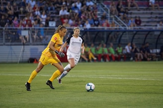 A soccer player in yellow goalkeeper attire is advancing with a soccer ball on a grassy field. Another player in white is running behind. A blurred crowd is visible in the stands, alongside other players seated on the bench.