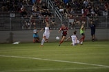A competitive soccer match is taking place with players in action on the field. Two players in white uniforms are being challenged by an opponent in a red and black uniform. The crowd in the background is seated in bleachers, watching the intense play. The field is well-lit, and a ball is in motion near the players.