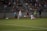 A competitive soccer match is taking place with players in action on the field. Two players in white uniforms are being challenged by an opponent in a red and black uniform. The crowd in the background is seated in bleachers, watching the intense play. The field is well-lit, and a ball is in motion near the players.