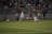 A competitive soccer match is taking place with players in action on the field. Two players in white uniforms are being challenged by an opponent in a red and black uniform. The crowd in the background is seated in bleachers, watching the intense play. The field is well-lit, and a ball is in motion near the players.