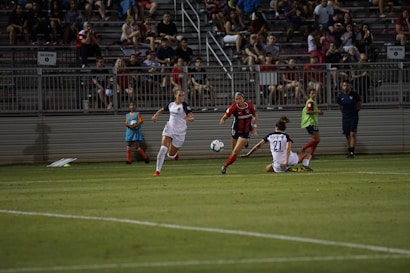 A competitive soccer match is taking place with players in action on the field. Two players in white uniforms are being challenged by an opponent in a red and black uniform. The crowd in the background is seated in bleachers, watching the intense play. The field is well-lit, and a ball is in motion near the players.