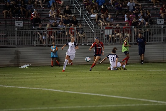 A competitive soccer match is taking place with players in action on the field. Two players in white uniforms are being challenged by an opponent in a red and black uniform. The crowd in the background is seated in bleachers, watching the intense play. The field is well-lit, and a ball is in motion near the players.