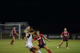 A women's soccer match is taking place at night on a well-lit field. Two players, one in a red and blue uniform and another in a white uniform, are intensely competing for the ball. In the background, more players in similar red and blue uniforms are visible, with goalposts and faint signage along the sidelines.