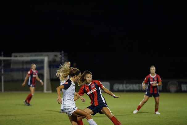 A women's soccer match is taking place at night on a well-lit field. Two players, one in a red and blue uniform and another in a white uniform, are intensely competing for the ball. In the background, more players in similar red and blue uniforms are visible, with goalposts and faint signage along the sidelines.