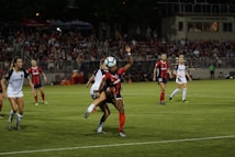 A soccer match with players on the field during an evening game. One player in a dark jersey is jumping to hit the ball, while others in white jerseys and dark shorts are positioned around. The stadium is filled with spectators watching the game, creating a lively atmosphere.