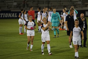 A group of female soccer players in white uniforms are gathered on a soccer field, some showing expressions of disappointment or exhaustion. A coach and a few other staff members are also present. The players appear to be walking off the field, and the environment suggests a professional or competitive match setting, with part of a stadium visible in the background.