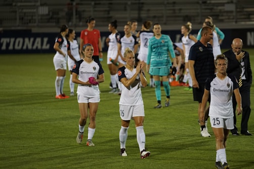 A group of female soccer players in white uniforms are gathered on a soccer field, some showing expressions of disappointment or exhaustion. A coach and a few other staff members are also present. The players appear to be walking off the field, and the environment suggests a professional or competitive match setting, with part of a stadium visible in the background.