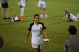 A soccer player in a white uniform with the number 10 is walking on a grassy field, smiling. Other players are visible in the background, some standing and some stretching on the grass. Several water bottles and bags are on the ground.