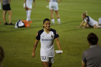 A soccer player in a white uniform with the number 10 is walking on a grassy field, smiling. Other players are visible in the background, some standing and some stretching on the grass. Several water bottles and bags are on the ground.