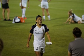A soccer player in a white uniform with the number 10 is walking on a grassy field, smiling. Other players are visible in the background, some standing and some stretching on the grass. Several water bottles and bags are on the ground.