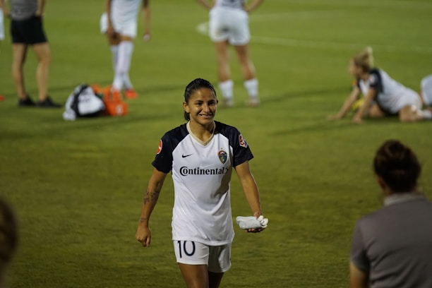 A soccer player in a white uniform with the number 10 is walking on a grassy field, smiling. Other players are visible in the background, some standing and some stretching on the grass. Several water bottles and bags are on the ground.