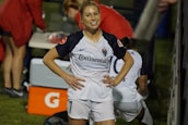 A female soccer player with a ponytail stands smiling with her hands on her hips, wearing a white and navy soccer uniform with the number 7. In the background, other team members are visible, with a table and equipment in the scene.