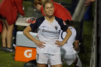 A female soccer player with a ponytail stands smiling with her hands on her hips, wearing a white and navy soccer uniform with the number 7. In the background, other team members are visible, with a table and equipment in the scene.