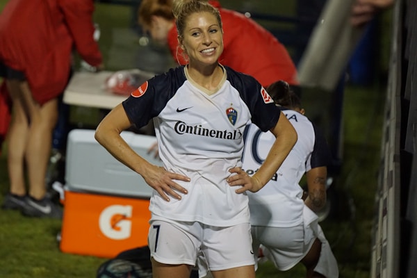 A female soccer player with a ponytail stands smiling with her hands on her hips, wearing a white and navy soccer uniform with the number 7. In the background, other team members are visible, with a table and equipment in the scene.