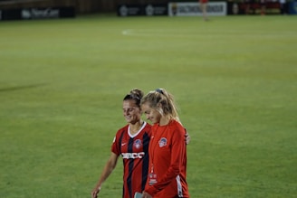 Two individuals walk arm-in-arm on a soccer field. One wears a red and black striped sports jersey, while the other is dressed in a solid red outfit with a staff badge. Both appear to be smiling, conveying a sense of camaraderie and friendliness.