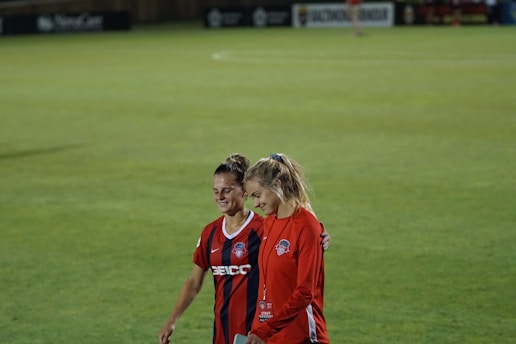 Two individuals walk arm-in-arm on a soccer field. One wears a red and black striped sports jersey, while the other is dressed in a solid red outfit with a staff badge. Both appear to be smiling, conveying a sense of camaraderie and friendliness.