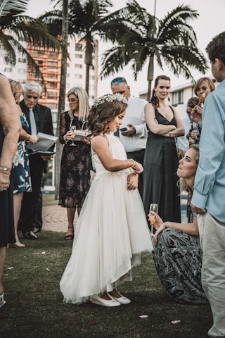 A young girl in a white dress with a floral crown stands among a group of elegantly dressed adults holding drinks, likely at an outdoor social event or celebration. In the background, palm trees and modern buildings are visible.