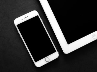 A smartphone and a tablet are placed on a dark surface, both devices have a sleek, modern design with a black screen and white casing. The smartphone is positioned on the left, slightly above the tablet, which is oriented horizontally.