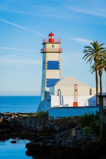 A lighthouse with a red top and blue and white checkered patterns stands near a rocky shoreline with the ocean in the background. Next to the lighthouse, there is a white building with an orange pole structure on the side. Palm trees are also visible near the building.