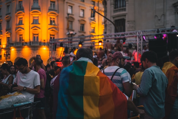 A crowd gathers in an outdoor urban setting, possibly a parade or festival, with attendees draped in rainbow flags. The scene is set during evening time, with a prominent building in the background illuminated by warm, yellow lights. People are engaged in conversations and taking photos, creating a lively atmosphere.