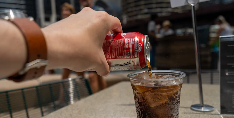 A hand pours soda from a red can into a plastic cup filled with ice, set on an outdoor table. The background shows blurred figures of people and buildings, indicating a public setting.
