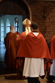 Several people dressed in religious robes walk down a hallway. One individual is carrying a bishop's crozier and another is wearing a mitre. The hallway has arched brick walls and windows on the left side, providing natural light.