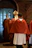 Several people dressed in religious robes walk down a hallway. One individual is carrying a bishop's crozier and another is wearing a mitre. The hallway has arched brick walls and windows on the left side, providing natural light.