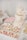 An elegant dessert table features a two-tiered cake adorned with delicate pink and white floral decorations. The cake has 'Engaged' written in gold, symbolizing a celebration. Surrounding it are an assortment of cupcakes topped with pastel-colored frosting, pink and white macarons, and custom sugar cookies with inscriptions. Decorative elements like lace and floral themes add to the sophistication and romantic atmosphere.