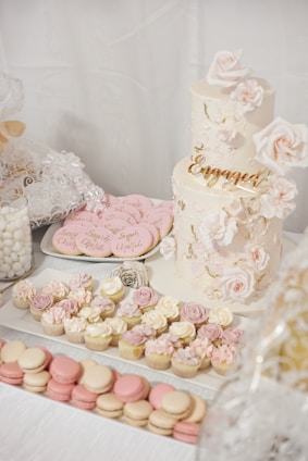 An elegant dessert table features a two-tiered cake adorned with delicate pink and white floral decorations. The cake has 'Engaged' written in gold, symbolizing a celebration. Surrounding it are an assortment of cupcakes topped with pastel-colored frosting, pink and white macarons, and custom sugar cookies with inscriptions. Decorative elements like lace and floral themes add to the sophistication and romantic atmosphere.