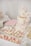 An elegant dessert table features a two-tiered cake adorned with delicate pink and white floral decorations. The cake has 'Engaged' written in gold, symbolizing a celebration. Surrounding it are an assortment of cupcakes topped with pastel-colored frosting, pink and white macarons, and custom sugar cookies with inscriptions. Decorative elements like lace and floral themes add to the sophistication and romantic atmosphere.