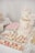 An elegant dessert table features a two-tiered cake adorned with delicate pink and white floral decorations. The cake has 'Engaged' written in gold, symbolizing a celebration. Surrounding it are an assortment of cupcakes topped with pastel-colored frosting, pink and white macarons, and custom sugar cookies with inscriptions. Decorative elements like lace and floral themes add to the sophistication and romantic atmosphere.