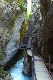 A narrow canyon with steep rock walls, featuring a wooden walkway suspended along the sides. A vibrant blue river flows below, cutting through the rocky terrain. Sparse greenery clings to the rocks, adding touches of color to the predominantly gray stone.