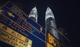 A large blue street sign with multiple directional arrows and text in both English and Malay is prominently visible. Behind the sign, two tall, illuminated twin towers stand against a dark night sky.