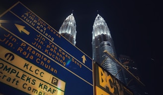 A large blue street sign with multiple directional arrows and text in both English and Malay is prominently visible. Behind the sign, two tall, illuminated twin towers stand against a dark night sky.