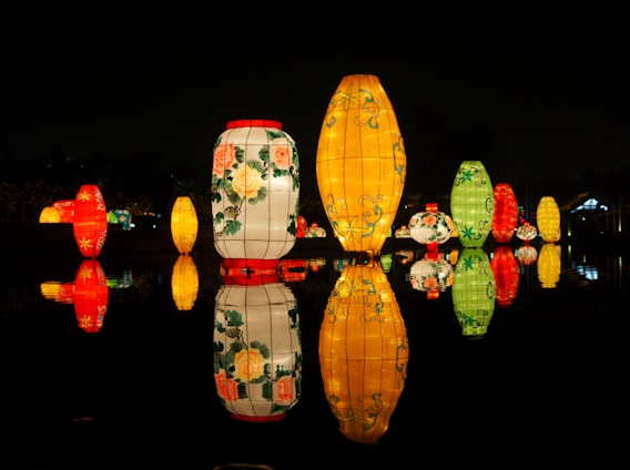 Colorful lanterns of various shapes and sizes float on water, their vibrant hues reflecting on the dark, still surface. Each lantern displays intricate patterns, including floral and geometric designs, and emits a warm, inviting glow against the night sky.
